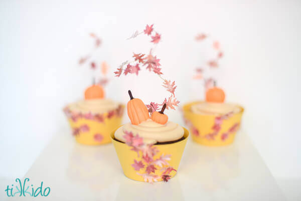 Three cupcakes topped with gum paste pumpkins and fall leaf cupcake toppers.