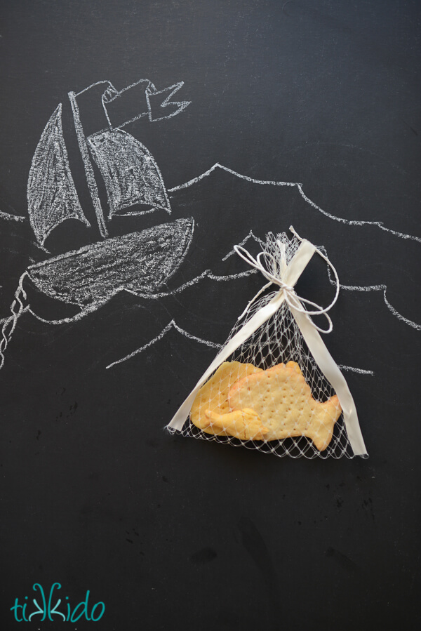 Homemade goldfish crackers in a net bag on a black chalkboard drawn with a boat and ocean waves.
