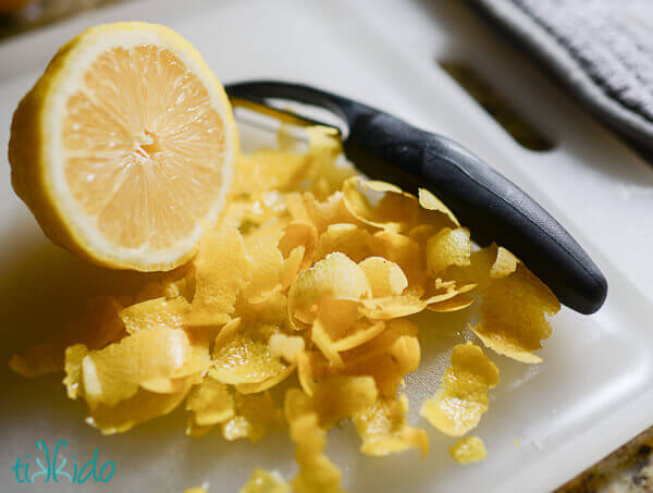 Lots of lemon peel next to a sliced lemon and a vegetable peeler on a white cutting board.
