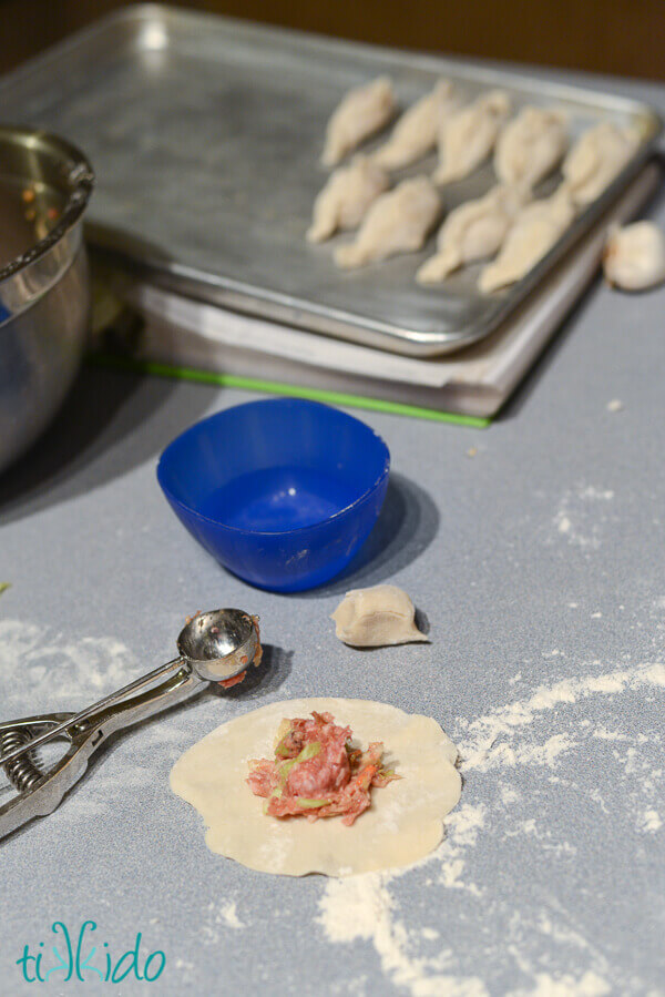Homemade Chinese dumpling wrapper being filled with a pork mixture, next to a sheet pan with finished dumplings.