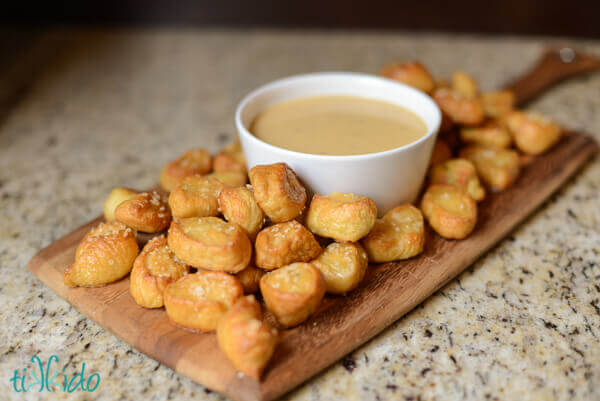White bowl of beer cheddar sauce surrounded by homemade pretzel bites, displayed on a wooden cutting board on a granite counter.