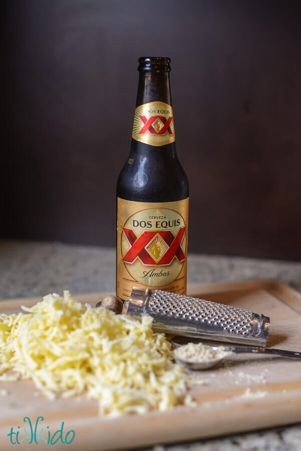 Bottle of Dos Equis beer, surrounded by a nutmeg grater, a heaping teaspoon of flour, and a pile of shredded white cheddar cheese.