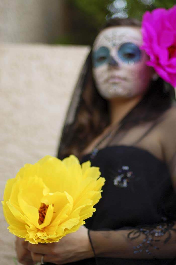 Woman in Dia de los Muertos makeup and a black dress holding a giant yellow tissue paper flower.