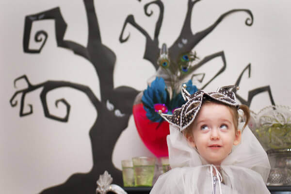 Little girl wearing a hooded tulle cape in front of a spooky tree background.