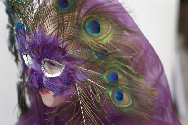 Closeup of a hood of a purple tulle cape, worn by a young girl wearing a peacock feather masquerade mask.