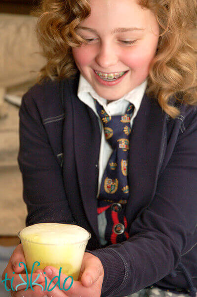 Girl dressed in Harry Potter costume smiling at clear cup filled with foaming yellow liquid.