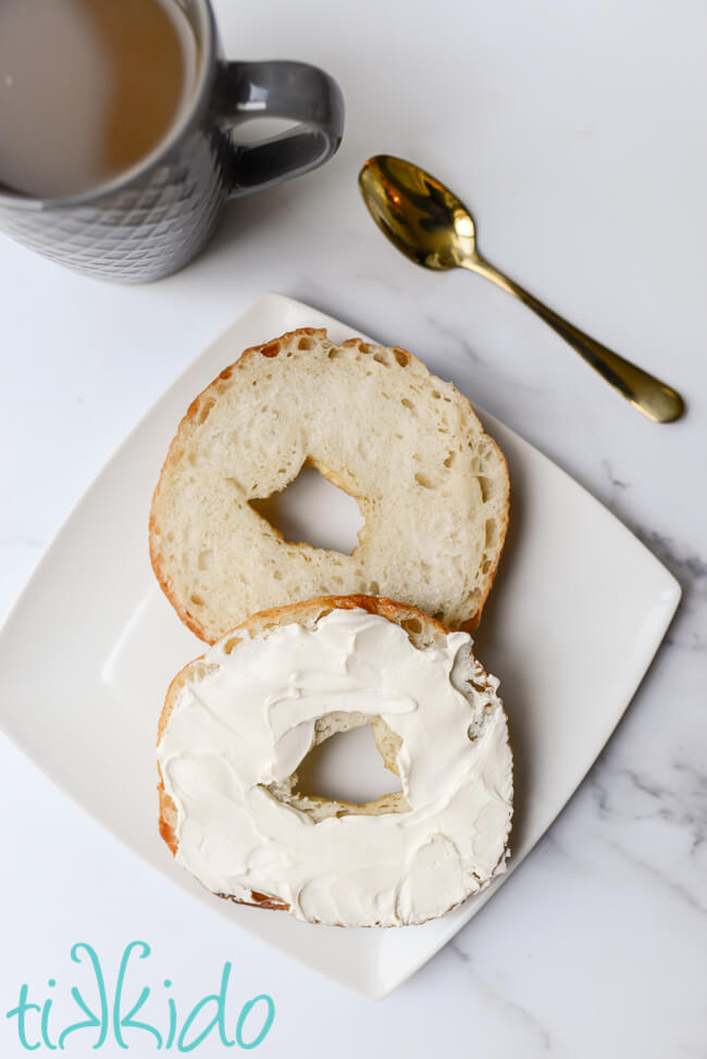 homemade bagel sliced in half and spread with cream cheese, next to a cup of coffee and a golden spoon.