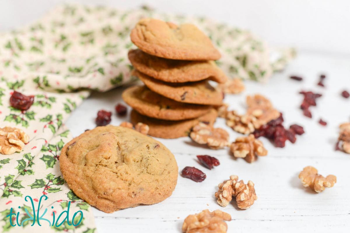 Cranberry Walnut Cookies Stack of Cranberry Walnut Cookies surrounded by walnut halves and dried cranberries and a holly print Christmas tea towel on a white wooden surface.