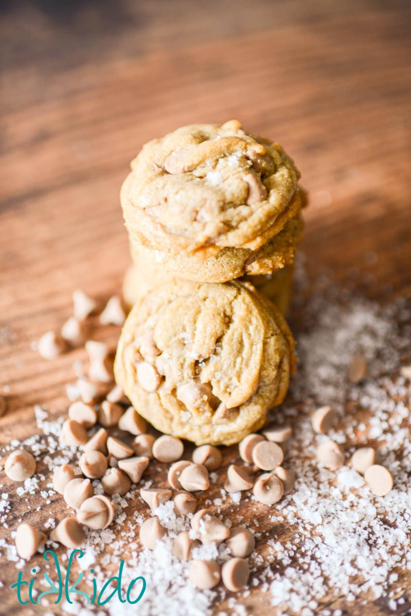 Salted Caramel Cookies stack of salted caramel cookies on a wooden surface, surrounded by salted caramel baking chips and flakes of sea salt.