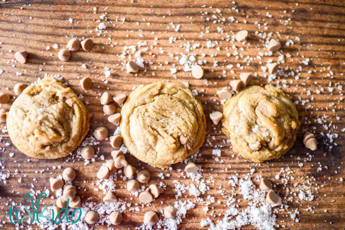 Salted Caramel Cookies Three salted caramel cookies in a row on a wooden surface, surrounded by salted caramel baking chips and flakes of sea salt.