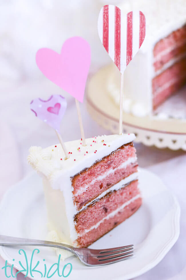Strawberry Cake Recipe Slice of strawberry cake on a white plate, topped with paper heart cake decorations.