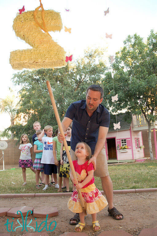 Small girl and her father hitting a yellow piñata shaped like the number 2.